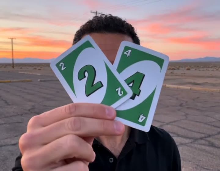 A frame from a generated 24 trend car video: a man holding up a green 2 card and a green 4 card in a parking lot at sunset